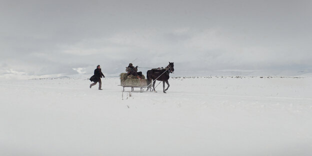 In einer verschneiten Landschaft wird ein Schlitten von einem Pferd gezogen, ein Mann läuft mit wehendem Mantel hinterher
